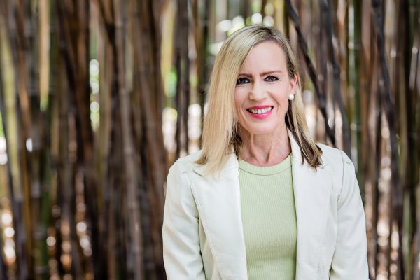 Portrait of Amy Walton. Amy is in a pale green blazer, smiling and standing in front of a bamboo grove. 